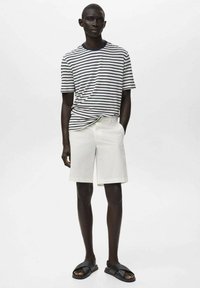 Randig marinblå och vit t-shirt med korta ärmar som matchas med vita shorts. Modellen bär svarta sandaler och visar upp en minimalistisk sommaroutfit.