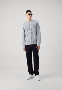 Sudadera azul claro con el texto "Racquet Club", combinada con jeans oscuros y zapatillas blancas. Modelo con gafas de sol negras se encuentra de pie frente a un fondo liso.