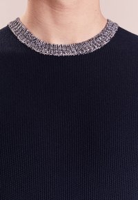 Gros plan sur une personne portant un pull en maille bleu marine foncé avec un col rond côtelé gris et blanc.