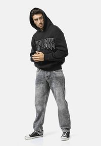 Schwarzer Kapuzenpullover mit fettem weißen Logo, kombiniert mit verblassten grauen Jeans und schwarzen High-Top-Sneakern. Das Modell posiert vor einem weißen Hintergrund.