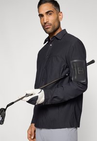 Μαύρο πουκάμισο με μακριά μανίκια, κλείσιμο με κουμπιά και τσέπη με φερμουάρ στον αριστερό μπράτσο. Άνδρας κρατάει μπαστούνι γκολφ, φοράει λευκό γάντι γκολφ.