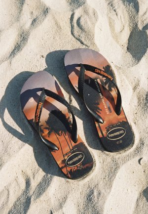 Par med svarte Havaianas flip-flops med palmetre-solnedgangsdesign som hviler på teksturert sand.