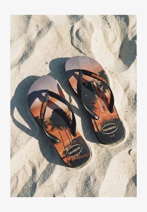 Paar schwarzer Havaianas-Zehentrenner mit Palmen-Sonnenuntergangs-Motiv, die auf strukturiertem Sand liegen.