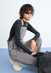 Gray denim overalls with adjustable straps, paired with a fitted black long-sleeve top and beige sneakers. Black bag beside.