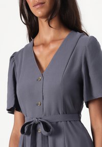 Abito grigio con scollatura a V, maniche a sbuffo corte e vita legata. Presenta dettagli con bottoni e una texture di tessuto liscia.