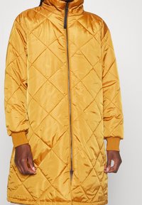 Manteau long matelassé jaune avec col montant, fermeture éclair à l'avant et poignets élastiques. Tissu lisse et brillant avec un motif losange.
