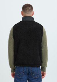 Gilet in finta pelliccia nera con colletto alto, indossato sopra un maglione verde. Presenta una consistenza morbida e una vestibilità rilassata, abbinato a jeans blu.