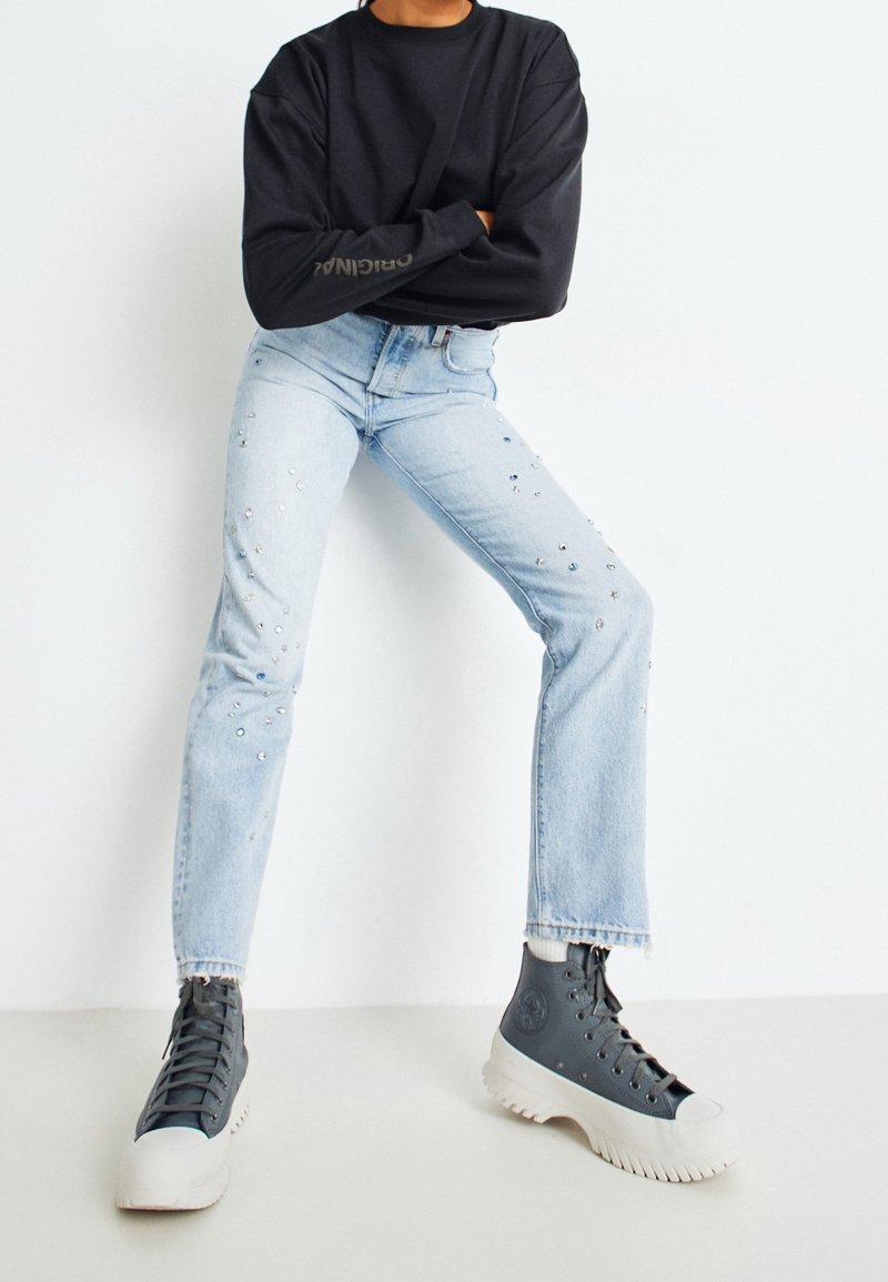 Top negro de manga larga combinado con jeans de mezclilla azul claro con adornos de rhinestones dispersos. El calzado son zapatillas deportivas altas grises con suelas blancas.