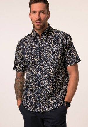 Uomo con barba e braccio tatuato che indossa una camicia blu navy a maniche corte con motivo floreale beige e pantaloni scuri, mani nelle tasche, su sfondo uniforme.