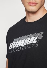 Must Black T-särk, millel on suur valge tekst "HUMMEL" erinevates stiilides ja orientatsioonides, ümmargune kaelus ja lühikesed varrukad.