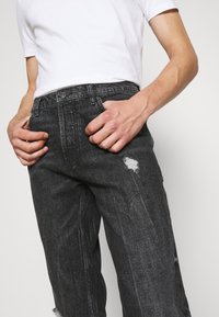Hombre vistiendo jeans rasgados negros y camiseta blanca con las manos cerca de los bolsillos frontales contra un fondo simple.