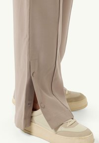 Light beige trousers with a slight sheen, featuring side slits secured by buttons, paired with off-white sneakers with a chunky sole.