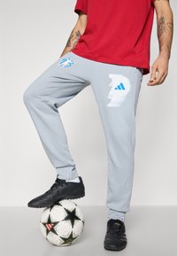 Svetlo šedé tepláky s voľným strihom, zdobené modrými grafickými dizajnmi a logom Adidas. Čierne futbalové topánky na vzorovanej lopte.