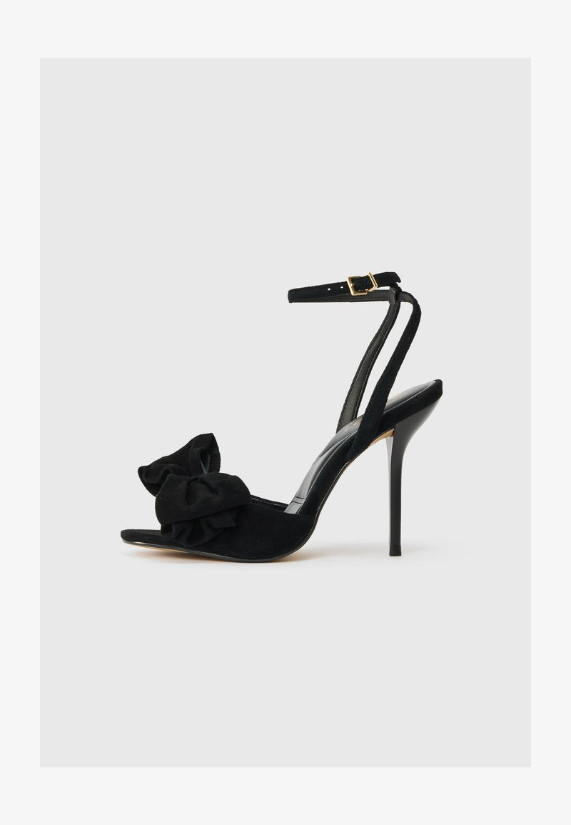 Sandale à talons hauts en daim noir avec une bride à la cheville, présentant un grand nœud décoratif au niveau des orteils et une semelle intérieure brillante. Design stilettos.