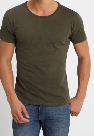 Homme portant un t-shirt ajusté à manches courtes couleur olive et un jean bleu, debout avec une main près de la poche, sur un fond uni.