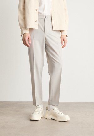 Osoba nosząca jasnobeżowe, dopasowane spodnie, białe skarpetki, masywne kremowe sneakersy oraz kremową zapinaną na guziki kurtkę, stojąca na szarej podłodze.