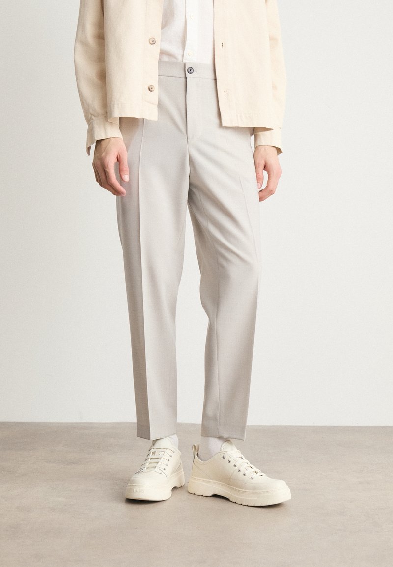 Person trägt hellbeige maßgeschneiderte Hose, weiße Socken, klobige cremefarbene Sneaker und eine cremefarbene, geknöpfte Jacke und steht auf einem grauen Boden.