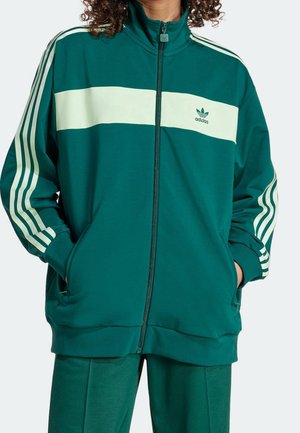 Persoon draagt een turquoise Adidas trainingsjack met witte strepen op de mouwen en borst, handen in de zakken, voorrits gesloten.