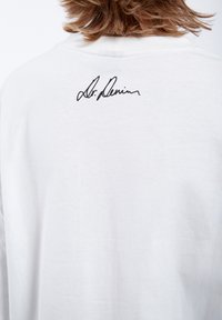 White cotton t-shirt with a crew neckline, featuring a black signature print on the upper back. Soft texture, relaxed fit design.