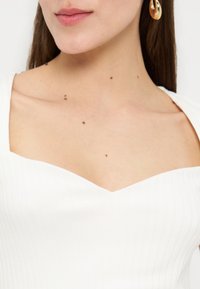 Γυναίκα που φοράει λευκό μπλουζάκι με ριμπ ύφανση και τετράγωνη λαιμόκοψη, με χρυσούς κρίκους στο αυτί, δείχνοντας τον λαιμό, τις κλείδες και ελιές στο δέρμα.