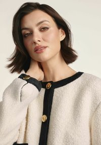 Kurtka polarowa w kremowym kolorze z czarnymi akcentami, z okrągłym dekoltem, złotymi guzikami i teksturowaną powierzchnią.