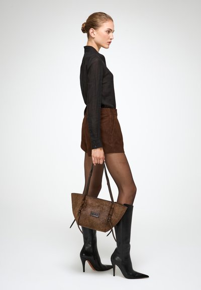 Chemise noire transparente à manches longues, shorts en suède marron, bottes noires hautes à talons aiguilles, et un sac à main marron texturé de forme triangulaire.