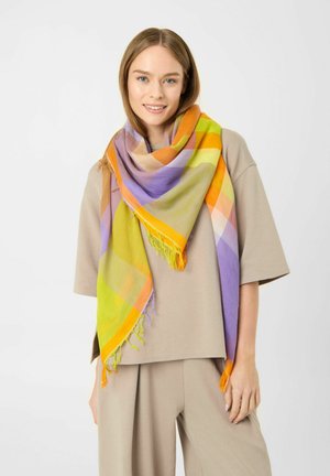 Junge Frau lächelt, trägt beigen oversized Pullover und Hose, mit großem buntem Karoschal in Orange, Gelb, Grün und Lila um den Hals gewickelt.