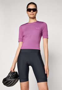 Maillot de cyclisme violet à manches courtes avec une coupe ajustée, associé à un short noir rembourré et un casque noir tenu dans la main gauche.