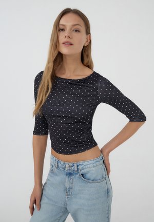 Camicia nera attillata con pois bianchi, maniche a tre quarti e scollatura a barca, abbinata a jeans in denim chiaro.
