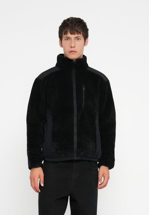 Black fleece jacket with high collar, zip closure, textured fabric, and solid black side panels. Worn with black jeans.