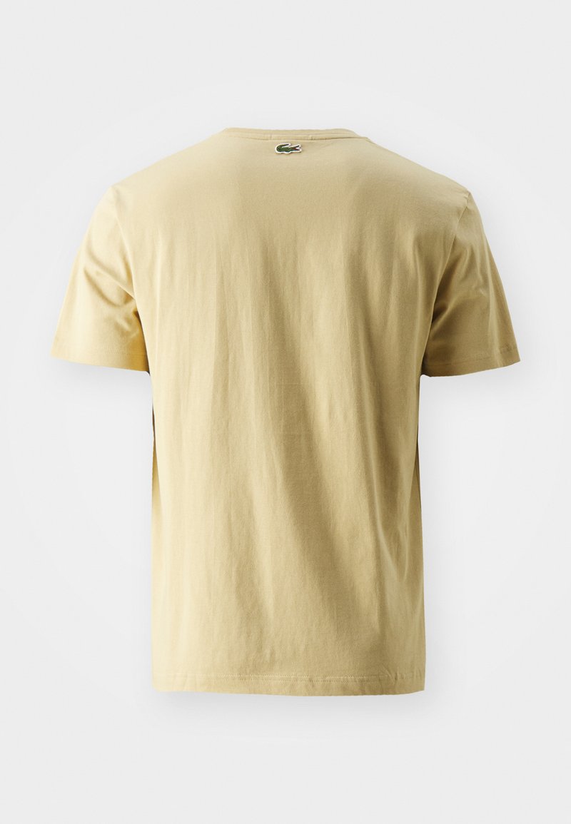 Camiseta de manga corta beige con un pequeño logo de cocodrilo verde centrado cerca del cuello en la parte de atrás.