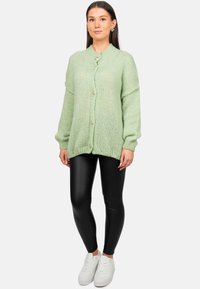 Cardigan lavorato a maglia verde chiaro con scollo rotondo, chiusura a bottoni e maniche oversize, abbinato a leggings neri e sneakers bianche.