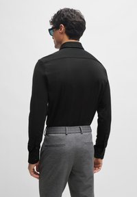 Schwarzes Langarmhemd mit geknöpften Manschetten, kombiniert mit grauen strukturierten Hosen mit flachem Bund und zwei Gesäßtaschen.