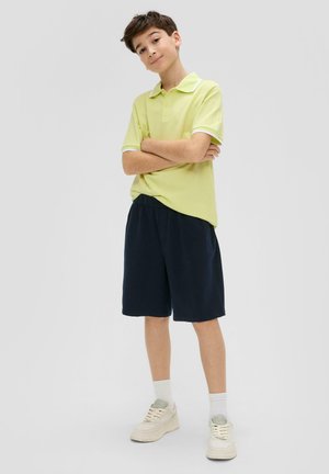 Polohemd in hellgelb mit weißem Kragen, kombiniert mit marineblauen Shorts. Die Schuhe sind hell und haben eine strukturierte Sohle. Das Model steht mit verschränkten Armen.