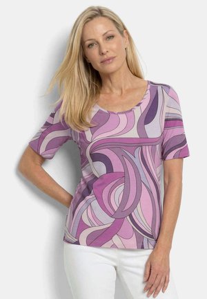 Femme aux longs cheveux blonds portant un haut à manches courtes violet et rose à motif abstrait et un pantalon blanc, debout avec une main sur la hanche.
