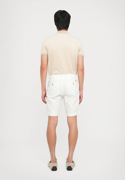 Beige teksturert polo-skjorte med korte ermer, kombinert med hvite knelengde shorts og lyse beige slip-on sko. Sett fra baksiden.