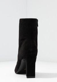 Bottine en daim noir avec un talon bloc haut, une texture lisse, une fermeture éclair latérale et un design à bout arrondi.