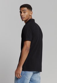 Zwarte polo met een kraag en korte mouwen. De achterzijde toont een gladde textuur en een ontspannen pasvorm. Gemaakt in combinatie met lichtblauwe denim jeans.