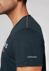 Mörkblå bomulls t-shirt med rund hals, med orange sömmar på axeln och en märkesetikett på vänster ärm.