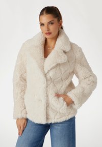 Kreminė dirbtinė pūkinė striukė su kvadratinio rašto, plati apykakle, priekinėmis kišenėmis ir švelniu, tekstūriniu paviršiumi. Derinama su mėlynais džinsais.