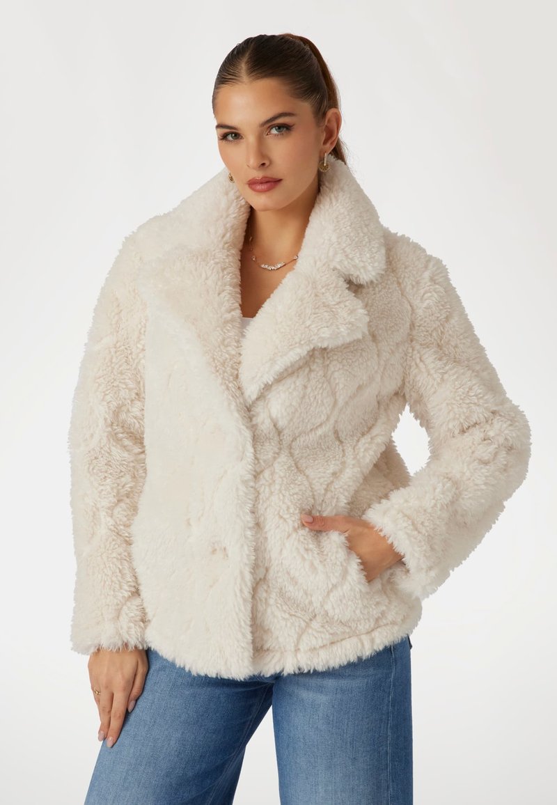 Kreminė dirbtinė pūkinė striukė su kvadratinio rašto, plati apykakle, priekinėmis kišenėmis ir švelniu, tekstūriniu paviršiumi. Derinama su mėlynais džinsais.