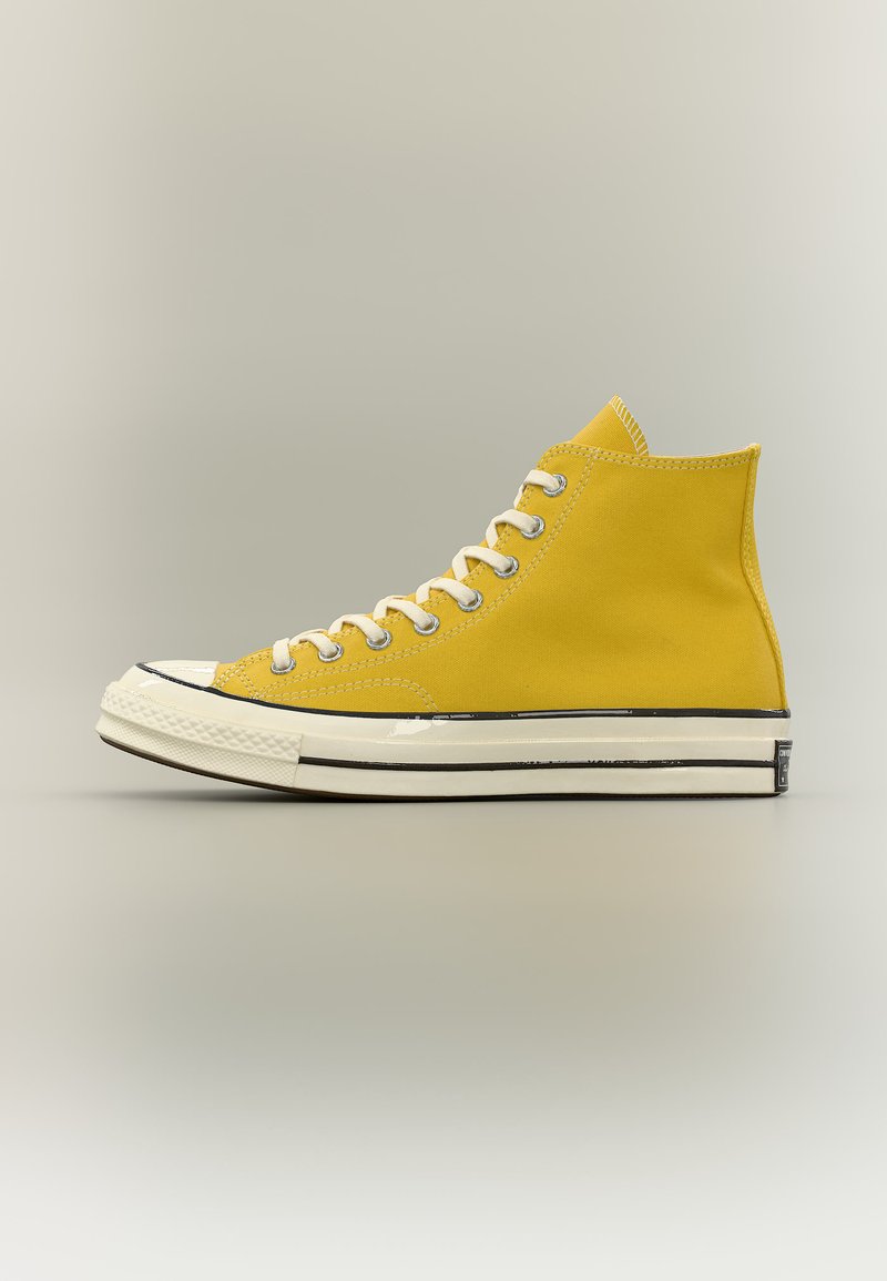 Baskets montantes en toile jaune avec lacets blancs et semelle blanche, bordure noire, et embout en caoutchouc sur un fond neutre uni.