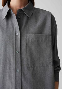 Close-up of a person wearing a gray button-up shirt with a chest pocket and rolled-up sleeve on a neutral background.
