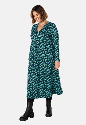 Langes schwarzes Kleid mit floralem Muster in Blau und Grün, mit V-Ausschnitt, langen Ärmeln und einem Bindeband in der Taille. Akzentuiert mit schwarzen Stiefeln.