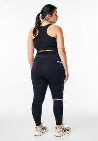 Czarny sportowy stanik i dopasowane czarne legginsy z białymi paskami akcentującymi. Tkaniny wyglądają na gładkie o elastycznej fakturze. Sneakersy widoczne poniżej.
