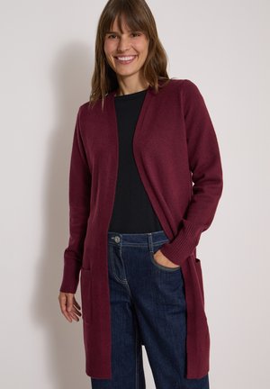 Femme souriante, portant un long cardigan bordeaux par-dessus un top noir et un jean bleu foncé, une main dans la poche, debout contre un mur uni.