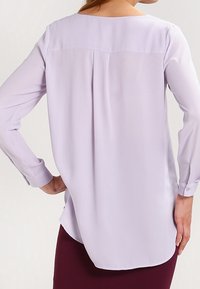 Blusón de lavanda claro con mangas largas, escote redondo y detalle de pliegue en la parte posterior. Tela suave y fluida; dobladillo curvado a la altura de la cintura.