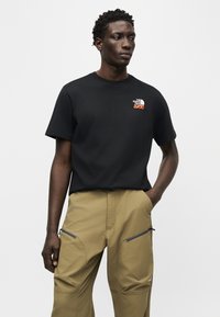 Άνδρας που φοράει μαύρο μπλουζάκι The North Face και μπεζ παντελόνι με τσέπες με φερμουάρ, στέκεται με το ένα χέρι στην τσέπη μπροστά σε απλό φόντο.