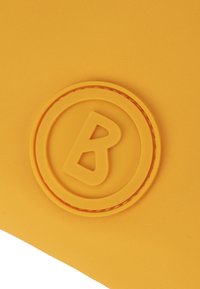 Žlutá taška s texturovaným povrchem, kulatým gumovým logem s písmenem "B" uprostřed a hladkým okrajem kolem loga.