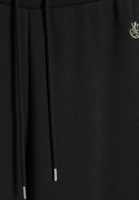 Czarne dresy z płaskim paskiem w talii i sznurkiem do regulacji. Posiadają złoty emblemat oraz strukturalną tkaninę. Miękka wykończenie z detalami w bocznych szwach.